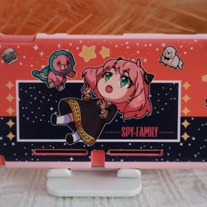 Puede incluir: Una funda rosa y roja para Nintendo Switch con Anya Forger de Spy x Family. La funda tiene un fondo negro y azul marino con estrellas y el texto "SPY x FAMILY". La funda también tiene ilustraciones de un oso, un perro y estrellas.