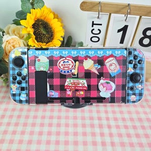May include: A Nintendo Switch console with a clear protective cover featuring a pink and black plaid pattern. The cover is decorated with various food-themed stickers and a blue bow design. The Joy-Con controllers have a blue plaid design.