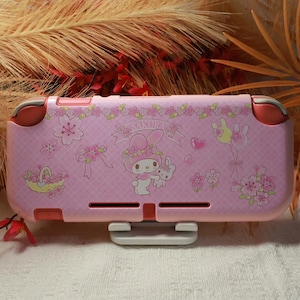 Op de afbeelding: Roze Nintendo Switch Lite hoesje met een My Melody ontwerp, kersenbloesems, harten en een konijn. De hoes heeft een lichtroze basis met rode accenten en een witte standaard.