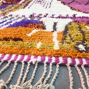 May include: Close-up of a colourful, textured rug with a landscape design. The rug features orange, purple, white, and pink hues, with a fringe detail along the bottom edge. The rug's design includes mountain-like shapes.