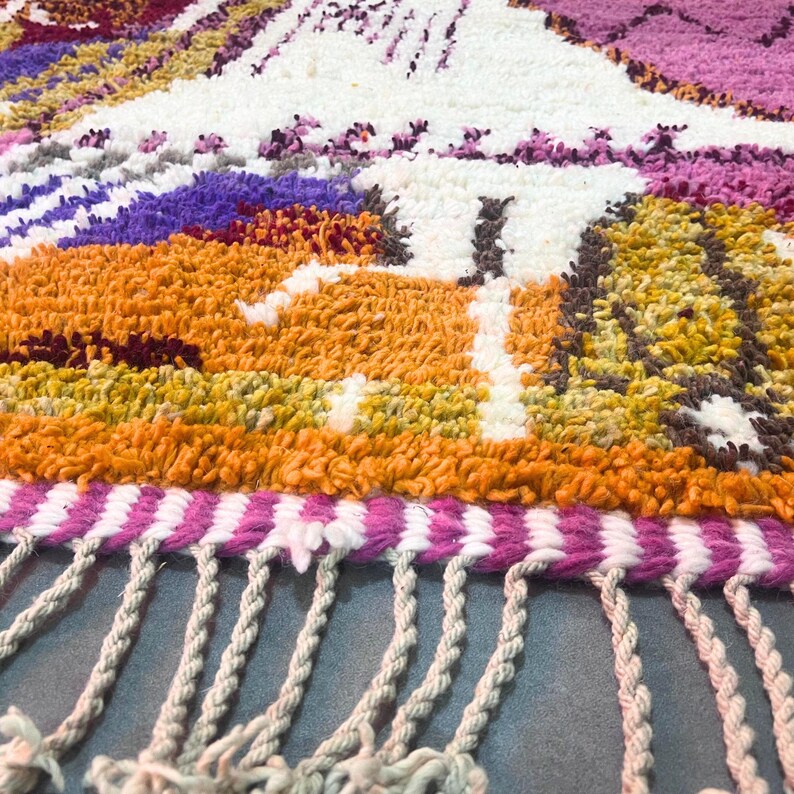 May include: Close-up of a colourful, textured rug with a landscape design. The rug features orange, purple, white, and pink hues, with a fringe detail along the bottom edge. The rug's design includes mountain-like shapes.