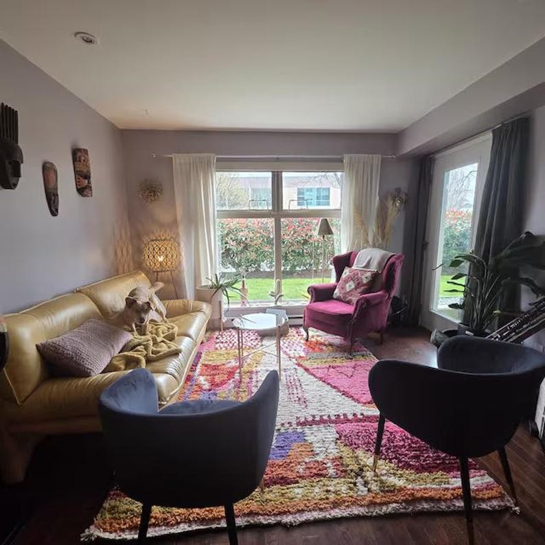 May include: A living room featuring a yellow leather sofa, a burgundy armchair, and two dark blue chairs. A colourful rug and decorative wall masks contribute to the room's eclectic style. A dog is on the sofa.