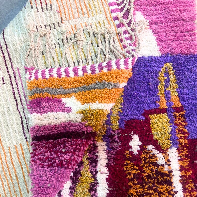 May include: A vibrant, textured rug with a patchwork design. The rug incorporates shades of pink, purple, orange, and white. It has a fringed edge and a geometric pattern. The rug's design is intricate and eye-catching.