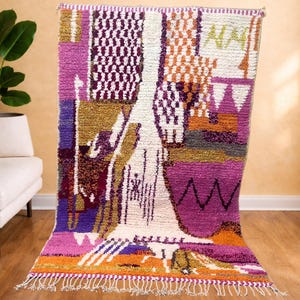 May include: A rectangular rug with a vibrant geometric pattern in shades of purple, pink, orange, white, and gold. The rug has a fringed edge and a textured, woven appearance, suitable for home decor, measuring approximately 200cm x 150cm.