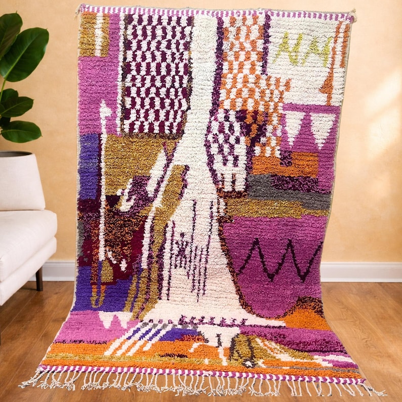 May include: A rectangular rug with a vibrant geometric pattern in shades of purple, pink, orange, white, and gold. The rug has a fringed edge and a textured, woven appearance, suitable for home decor, measuring approximately 200cm x 150cm.