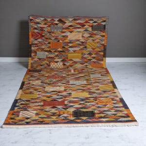 May include: A rectangular rug with a geometric pattern in shades of orange, yellow, red, and gray. The rug features a repeating design of triangles and squares, creating a patchwork effect. The rug has a fringed edge and is displayed on a white floor.