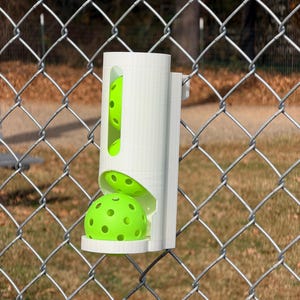 May include: A white, 3D-printed pickleball dispenser attached to a chain-link fence. The dispenser holds several neon green pickleballs with visible holes. The dispenser is designed to hold and dispense pickleballs for easy access during play.