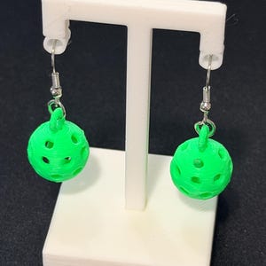 May include: A pair of bright green, spherical earrings with a perforated design. The earrings are suspended from silver-colored hooks and displayed on a white stand against a dark background. The earrings have a textured, 3D-printed appearance.
