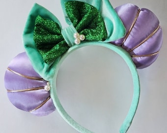 May include: A pair of green and purple headband ears with a green bow and pearl accents. The ears are shaped like seashells.