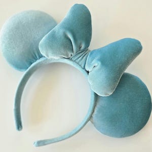 May include: A light blue velvet headband with two round ears and a bow. The headband is a solid color and has a soft, plush texture. The ears and bow are a slightly darker shade of blue.