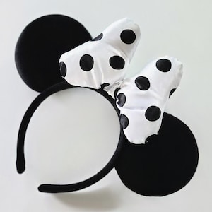 May include: Black headband with two large black circular ears and a white bow with black polka dots. The ears and headband are made of a soft, plush material. The bow is made of a silky fabric.