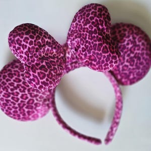 May include: A headband featuring two round, padded ears and a large central bow, all covered in a vibrant magenta and black leopard print fabric. The design includes a thin, flexible band that connects the two ears and the bow at the top center.