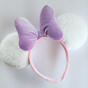 May include: Purple velvet bow headband with white fuzzy ears. The headband is pink.