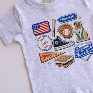 May include: A grey t-shirt with a variety of colorful patches, including a baseball, a baseball glove, a baseball bat, a s'more, a pennant that says "SUMMER '62", a dog wearing a baseball jersey, a pair of sneakers, and an American flag. The text "FOR-EV-ER" and "Legends Never Die" are also on the shirt.