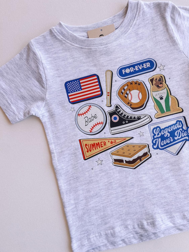 May include: A grey t-shirt with a variety of colorful patches, including a baseball, a baseball glove, a baseball bat, a s'more, a pennant that says "SUMMER '62", a dog wearing a baseball jersey, a pair of sneakers, and an American flag. The text "FOR-EV-ER" and "Legends Never Die" are also on the shirt.