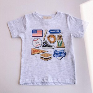 May include: A gray t-shirt with a variety of colorful patches, including a baseball, a baseball glove, a dog, a s'more, a sneaker, a baseball bat, a flag, and a pennant. The patches have text that reads "Babe", "FOR-EV-ER", "SUMMER '62", and "Legends Never Die!"
