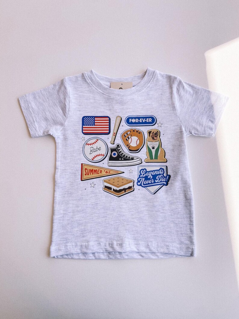 May include: A gray t-shirt with a variety of colorful patches, including a baseball, a baseball glove, a dog, a s'more, a sneaker, a baseball bat, a flag, and a pennant. The patches have text that reads "Babe", "FOR-EV-ER", "SUMMER '62", and "Legends Never Die!"