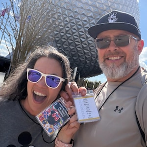 May include: A selfie of two people smiling in front of a large, geometric sphere. One person holds a lanyard with a "Drinking Around the World Passport" and a Mickey Mouse design. The other person holds a lanyard with a list of countries.