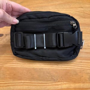 Puede incluir: Ri&ntilde;onera negra con cremallera superior y correa negra ajustable con hebilla de pl&aacute;stico. La ri&ntilde;onera est&aacute; sujeta por una mano y sobre una superficie de madera. Accesorio vers&aacute;til para llevar lo esencial.