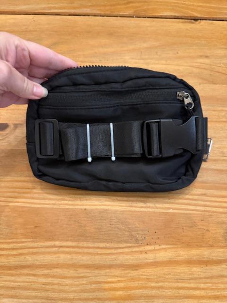 Puede incluir: Ri&ntilde;onera negra con cremallera superior y correa negra ajustable con hebilla de pl&aacute;stico. La ri&ntilde;onera est&aacute; sujeta por una mano y sobre una superficie de madera. Accesorio vers&aacute;til para llevar lo esencial.