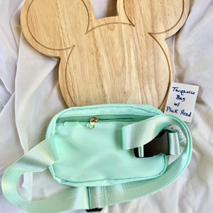 May include: A light turquoise belt bag with a zippered closure, a matching adjustable strap, and a black plastic buckle. A wooden board shaped like a mouse head is positioned behind the bag. A white tag reads Turquoise Bag w/ Pink Head.