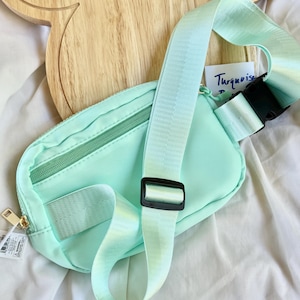 May include: A mint green belt bag featuring a zippered main compartment, an adjustable woven strap with a black plastic buckle, and a black plastic slider. A small white tag with the handwritten text Turquoise is visible near the strap.