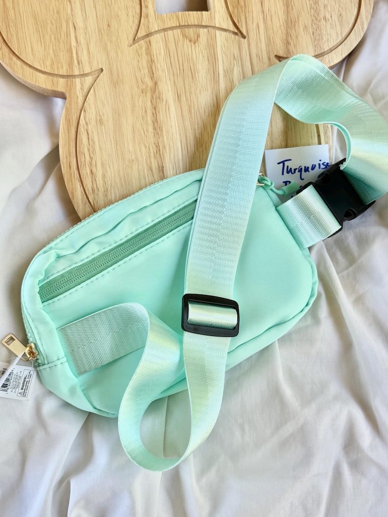 May include: A mint green belt bag featuring a zippered main compartment, an adjustable woven strap with a black plastic buckle, and a black plastic slider. A small white tag with the handwritten text Turquoise is visible near the strap.