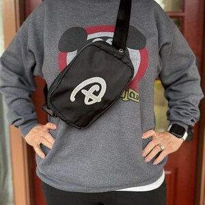 Puede incluir: Bolso bandolera negro con un logotipo plateado con purpurina. El bolso tiene una correa negra ajustable y cierre de cremallera. La persona lleva una sudadera gris con un dise&ntilde;o de Mickey Mouse.
