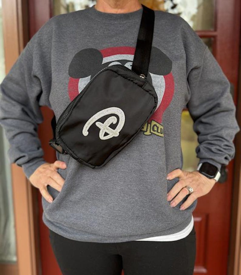 Puede incluir: Bolso bandolera negro con un logotipo plateado con purpurina. El bolso tiene una correa negra ajustable y cierre de cremallera. La persona lleva una sudadera gris con un dise&ntilde;o de Mickey Mouse.