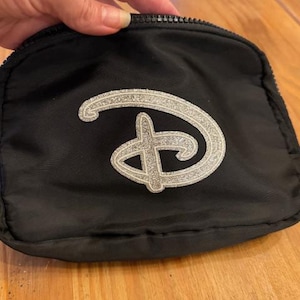 Puede incluir: Peque&ntilde;o neceser negro con cremallera y logotipo plateado brillante. El neceser es rectangular y parece estar hecho de tela resistente. El logotipo plateado es una letra "D" estilizada.