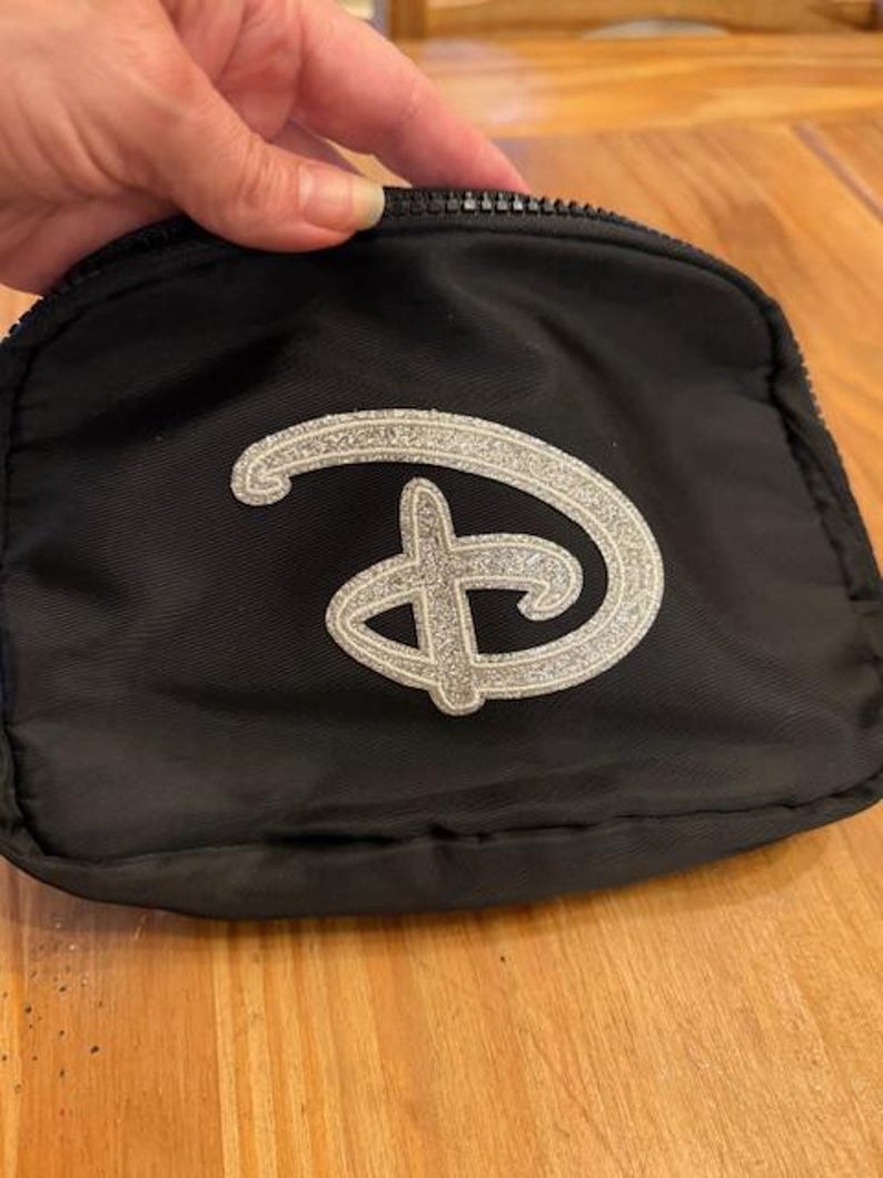 Puede incluir: Peque&ntilde;o neceser negro con cremallera y logotipo plateado brillante. El neceser es rectangular y parece estar hecho de tela resistente. El logotipo plateado es una letra "D" estilizada.