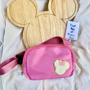 May include: A bright pink belt bag featuring a textured white Minnie Mouse head patch with a pink bow and gold trim. The bag has a matching pink adjustable strap with a black plastic buckle and a zipper closure. A small white tag reads Pink Bag w/ Minnie Head.