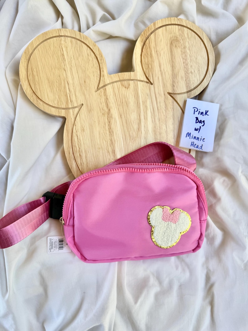 May include: A bright pink belt bag featuring a textured white Minnie Mouse head patch with a pink bow and gold trim. The bag has a matching pink adjustable strap with a black plastic buckle and a zipper closure. A small white tag reads Pink Bag w/ Minnie Head.