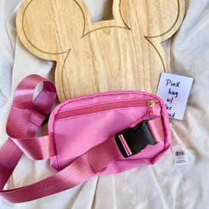 May include: A bright pink fanny pack with a matching adjustable strap, a black plastic buckle, and a gold zipper. Behind the bag is a light-colored wooden board shaped like a mouse head. A small white tag with the handwritten text Pink bag w/ blue bead is attached.