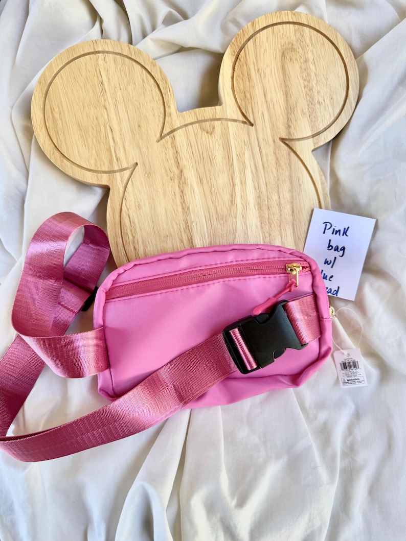 May include: A bright pink fanny pack with a matching adjustable strap, a black plastic buckle, and a gold zipper. Behind the bag is a light-colored wooden board shaped like a mouse head. A small white tag with the handwritten text Pink bag w/ blue bead is attached.