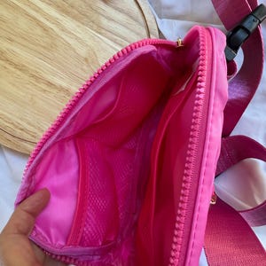 May include: A bright pink fanny pack with a matching pink zipper and an adjustable strap featuring a black plastic buckle. The interior is lined with pink fabric and includes a mesh pocket. A small white tag reads Minnie Head in blue ink.