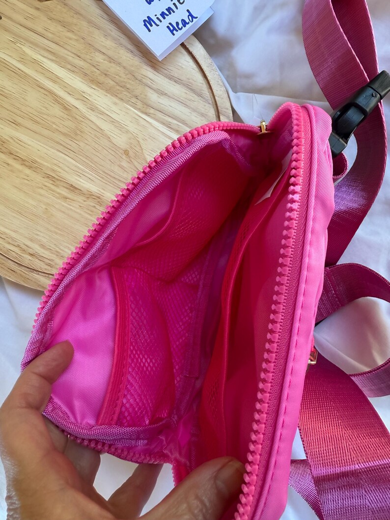 May include: A bright pink fanny pack with a matching pink zipper and an adjustable strap featuring a black plastic buckle. The interior is lined with pink fabric and includes a mesh pocket. A small white tag reads Minnie Head in blue ink.