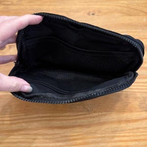 Puede incluir: Bolsa negra con cremallera abierta con m&uacute;ltiples compartimentos internos. La bolsa est&aacute; hecha de un material liso y mate y cuenta con un bolsillo de malla. La cremallera recorre la parte superior y los lados de la bolsa.