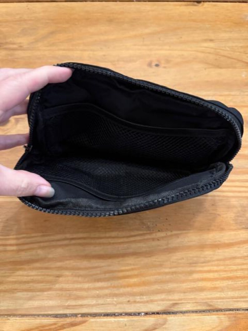 Puede incluir: Bolsa negra con cremallera abierta con m&uacute;ltiples compartimentos internos. La bolsa est&aacute; hecha de un material liso y mate y cuenta con un bolsillo de malla. La cremallera recorre la parte superior y los lados de la bolsa.
