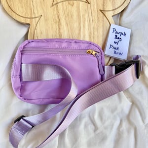 May include: A light purple fanny pack with a matching adjustable strap, a black plastic buckle, and a gold-tone zipper. A small white card is tucked behind the bag with the handwritten text: Purple Bag w/ Pink BOW.