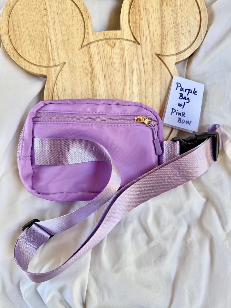 May include: A light purple fanny pack with a matching adjustable strap, a black plastic buckle, and a gold-tone zipper. A small white card is tucked behind the bag with the handwritten text: Purple Bag w/ Pink BOW.