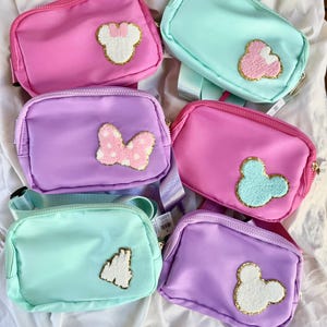 May include: Six rectangular belt bags in shades of pink, purple, and mint green. Each bag features a zippered closure and a textured, gold-trimmed patch on the front, including designs of mouse ears, a bow, and a castle silhouette.