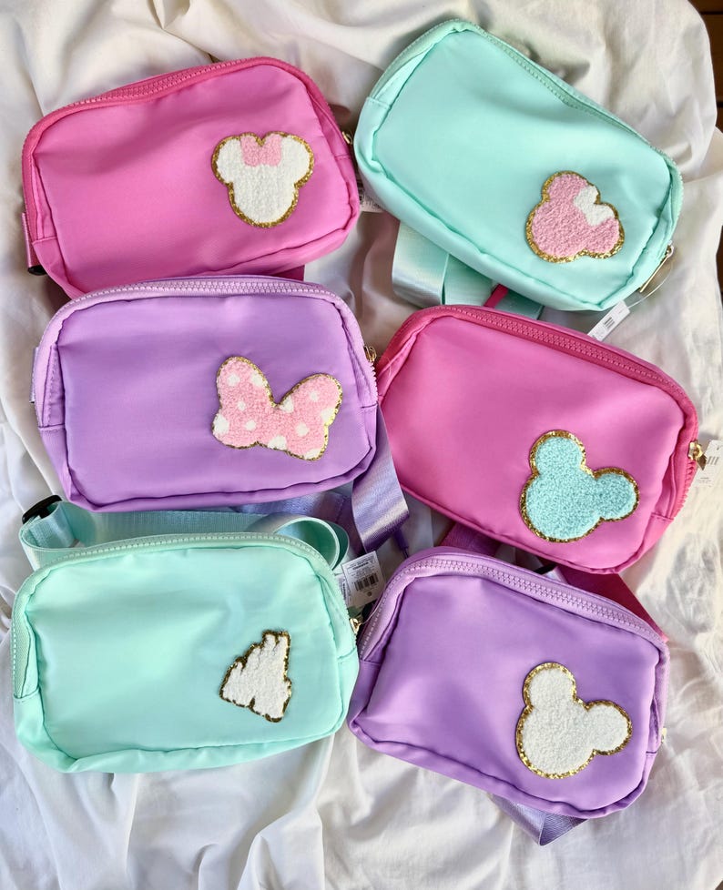 May include: Six rectangular belt bags in shades of pink, purple, and mint green. Each bag features a zippered closure and a textured, gold-trimmed patch on the front, including designs of mouse ears, a bow, and a castle silhouette.