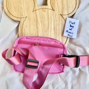May include: A bright pink fanny pack with a matching adjustable strap, a black plastic buckle, and a gold-tone zipper closure. A small white card is included with the handwritten text: Pink Bag w/ Minnie Head. The bag is positioned in front of a light-colored wooden board shaped like a mouse head.