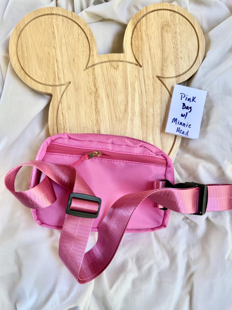 May include: A bright pink fanny pack with a matching adjustable strap, a black plastic buckle, and a gold-tone zipper closure. A small white card is included with the handwritten text: Pink Bag w/ Minnie Head. The bag is positioned in front of a light-colored wooden board shaped like a mouse head.