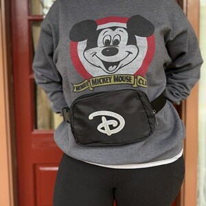 Puede incluir: Sudadera gris con un gr&aacute;fico de Mickey Mouse, un c&iacute;rculo rojo y el texto "MEMBER MICKEY MOUSE CLUB". Una ri&ntilde;onera negra con el logotipo de Disney con purpurina plateada se lleva alrededor de la cintura.