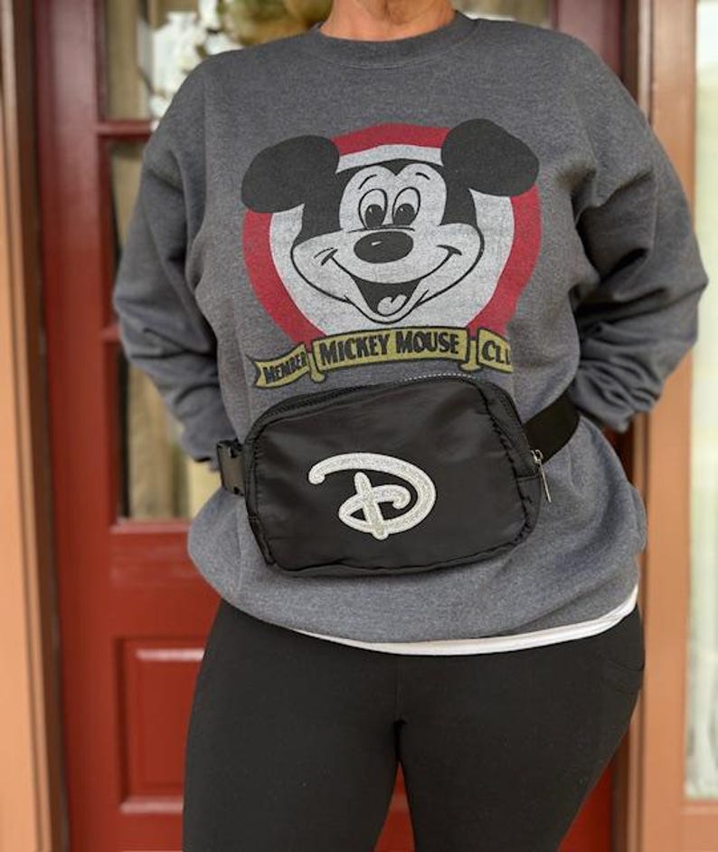 Puede incluir: Sudadera gris con un gr&aacute;fico de Mickey Mouse, un c&iacute;rculo rojo y el texto "MEMBER MICKEY MOUSE CLUB". Una ri&ntilde;onera negra con el logotipo de Disney con purpurina plateada se lleva alrededor de la cintura.