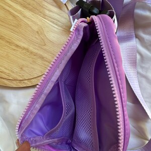 May include: A lavender belt bag with a matching adjustable strap and a black plastic buckle. The interior is lined in lavender fabric and features two mesh pockets on one side, secured by a thick lavender zipper that runs along the top edge.
