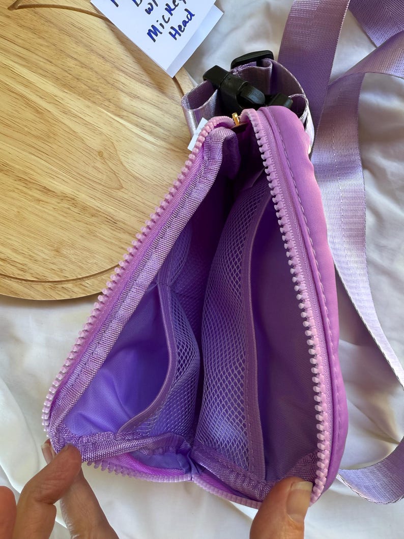May include: A lavender belt bag with a matching adjustable strap and a black plastic buckle. The interior is lined in lavender fabric and features two mesh pockets on one side, secured by a thick lavender zipper that runs along the top edge.