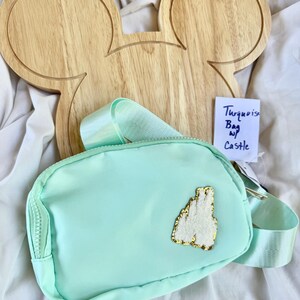 May include: A turquoise belt bag featuring a white textured castle patch with gold trim on the front. The bag has a matching turquoise adjustable strap and a zipper closure. A small white tag attached to the bag displays the handwritten text Turquoise Bag w/ Castle.
